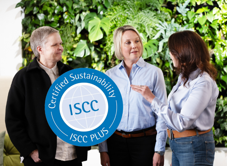 Three people discussing in front of a green plant wall. In front, the ISCC Plus logo.